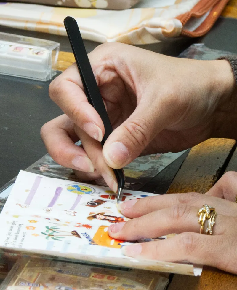 Unas manos colocando stickers ilustrados en una hoja con unas pinzas.